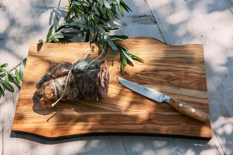 Wooden cutting board with cured meats and a knife on a light wooden floor.