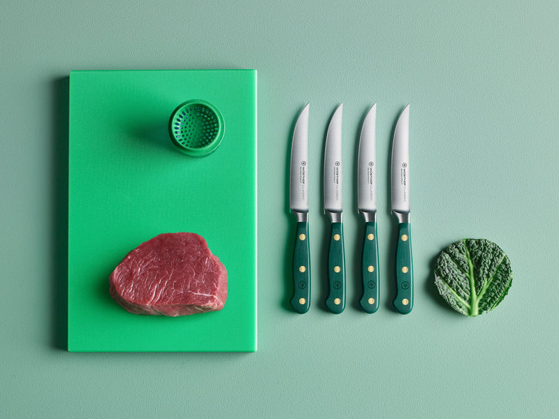 Green cutting board with a meat piece, spice container, four knives, and a cabbage leaf on a matching green surface.