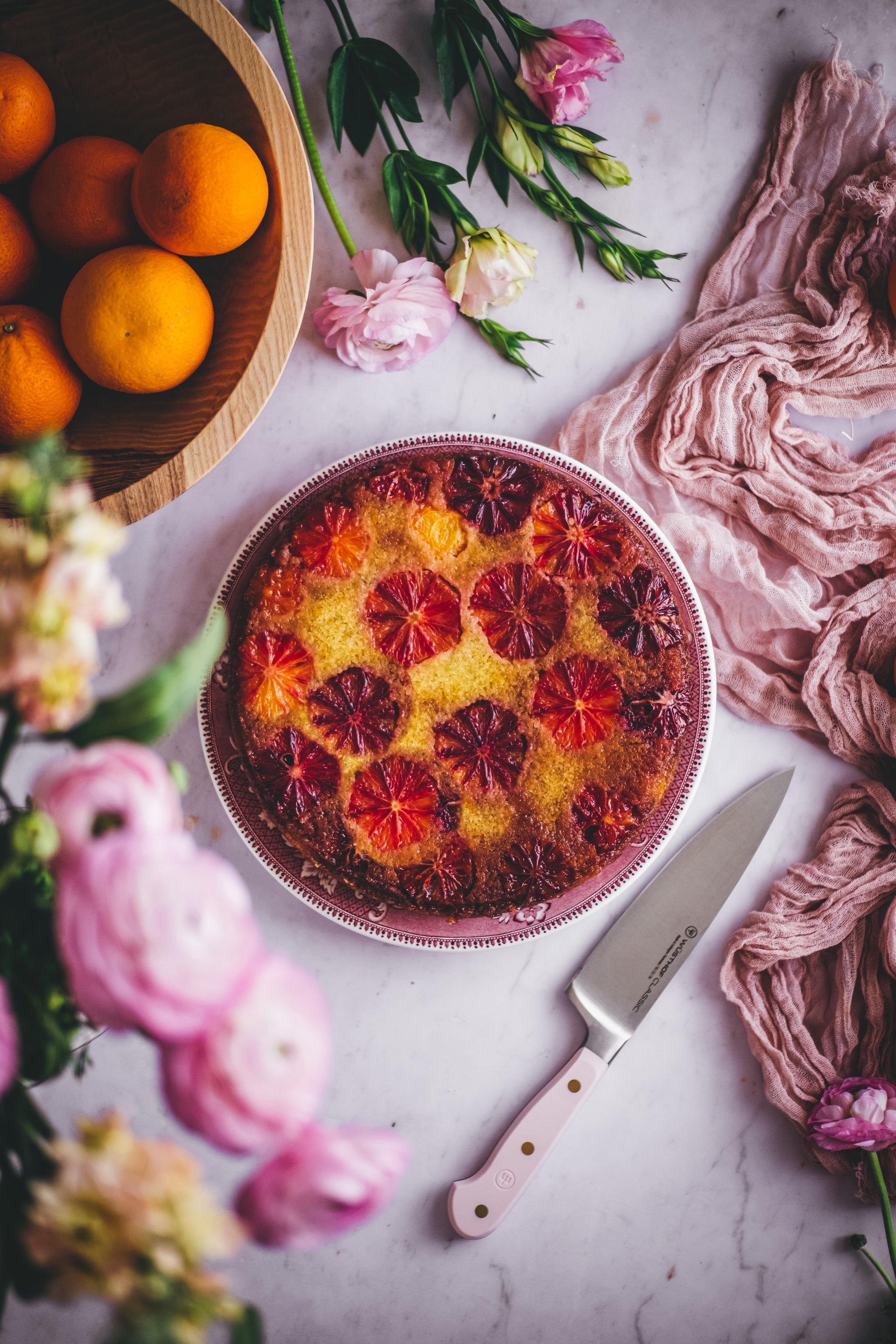 Decorative cake with colorful fruit on a table with flowers and a knife.