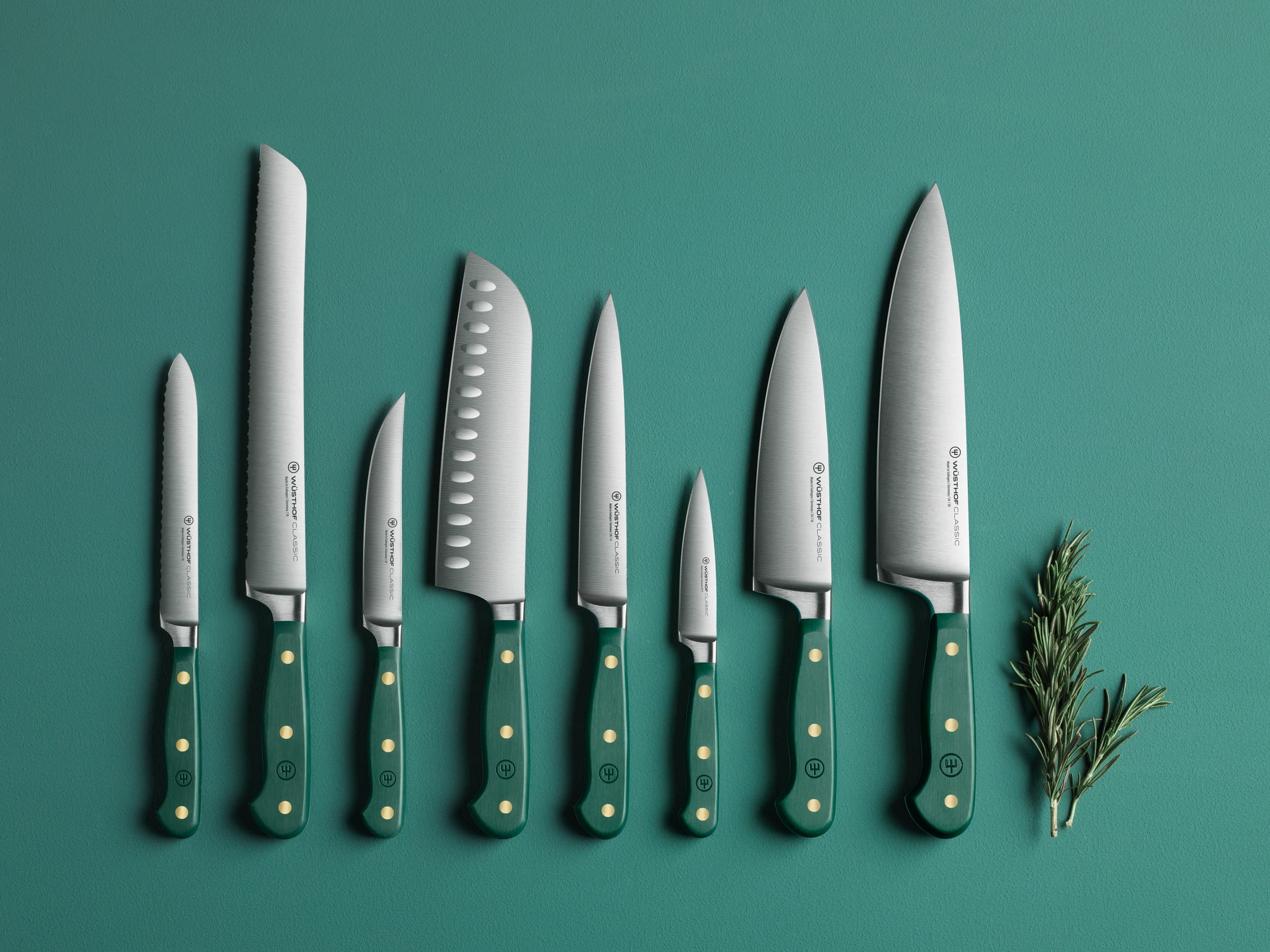 A set of eight kitchen knives with green handles arranged on a green surface, accompanied by sprigs of rosemary.