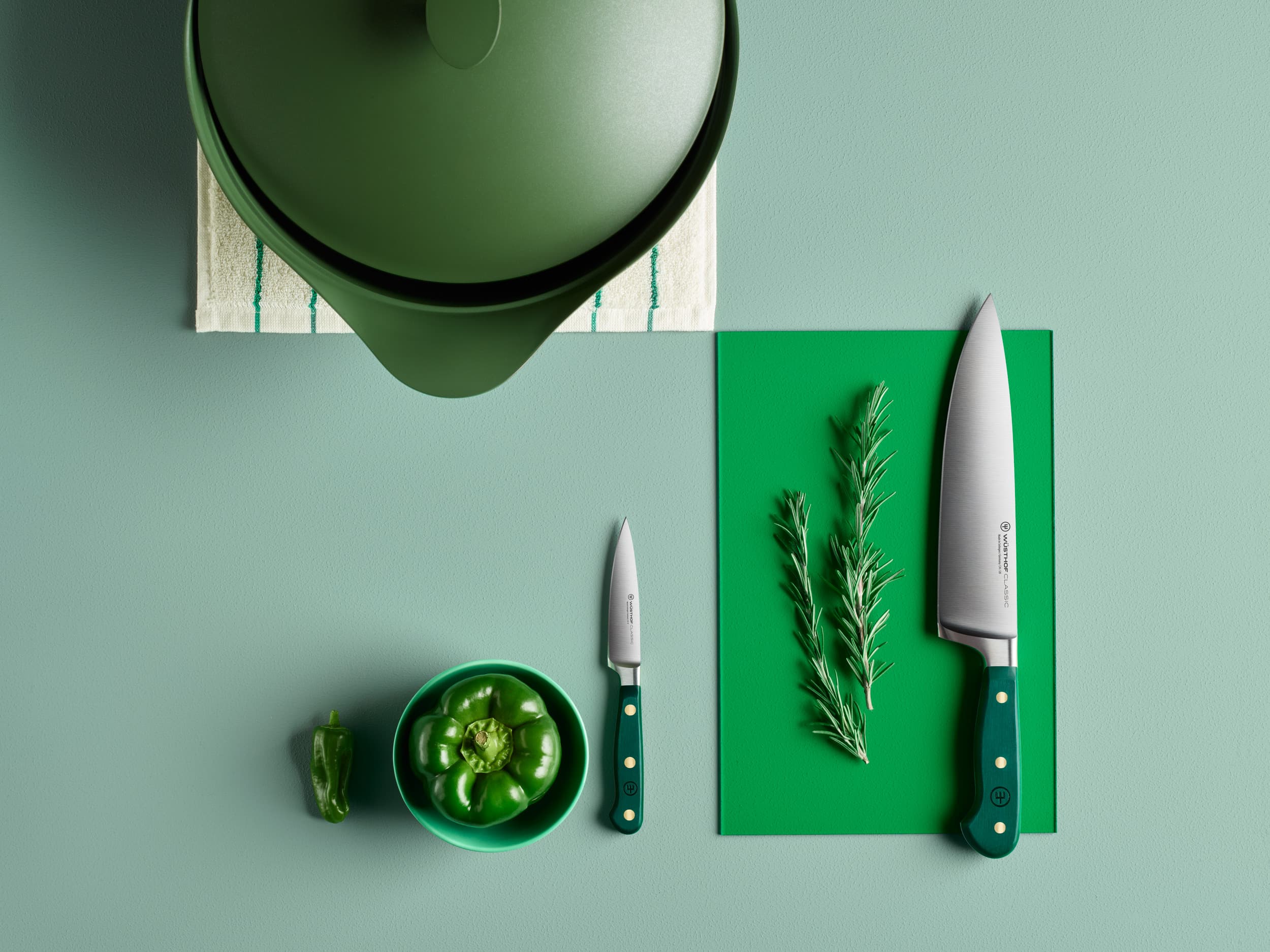 Top view of green cooking pot, knives, rosemary, green bell pepper, and cutting board on a green surface.