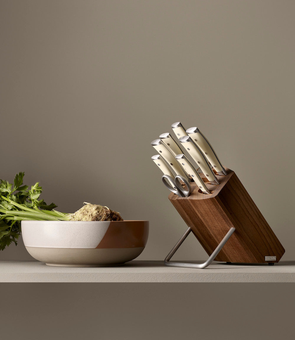 A kitchen knife block with cream-colored knives and a matching ceramic bowl on a shelf against a warm taupe background.