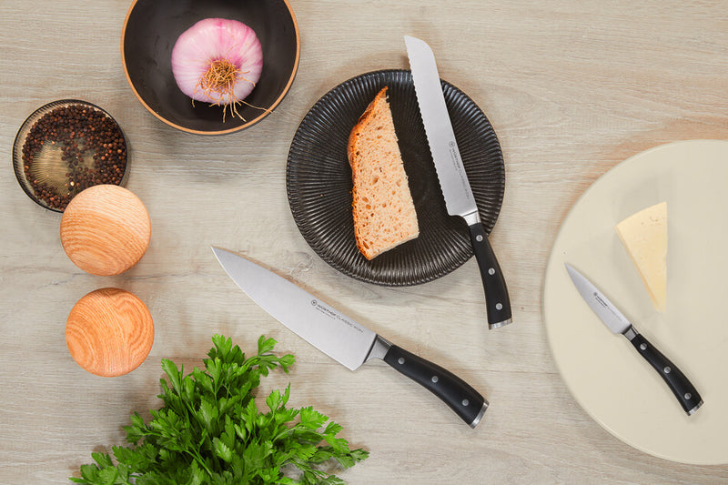 Classic Ikon 3-Piece Chef's Knife Set with three stainless steel knives featuring black handles, including a bread knife, chef’s knife, and paring knife displayed with food.