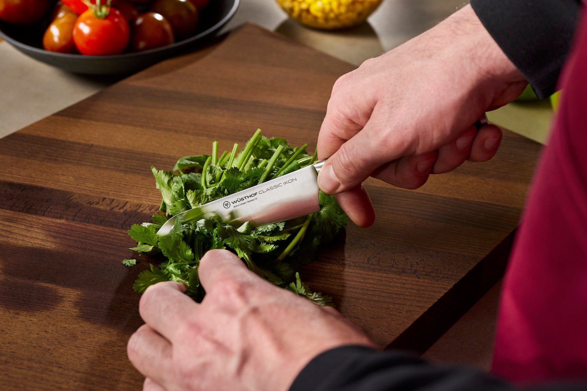 A close-up of a person's hands using an asian utility kitchen knife to chop green herbs on a dark wooden cutting board, with tomatoes visible in the background.