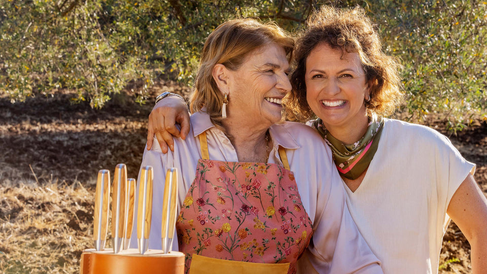 Two women standing outdoors with a rustic background, one wearing an apron.